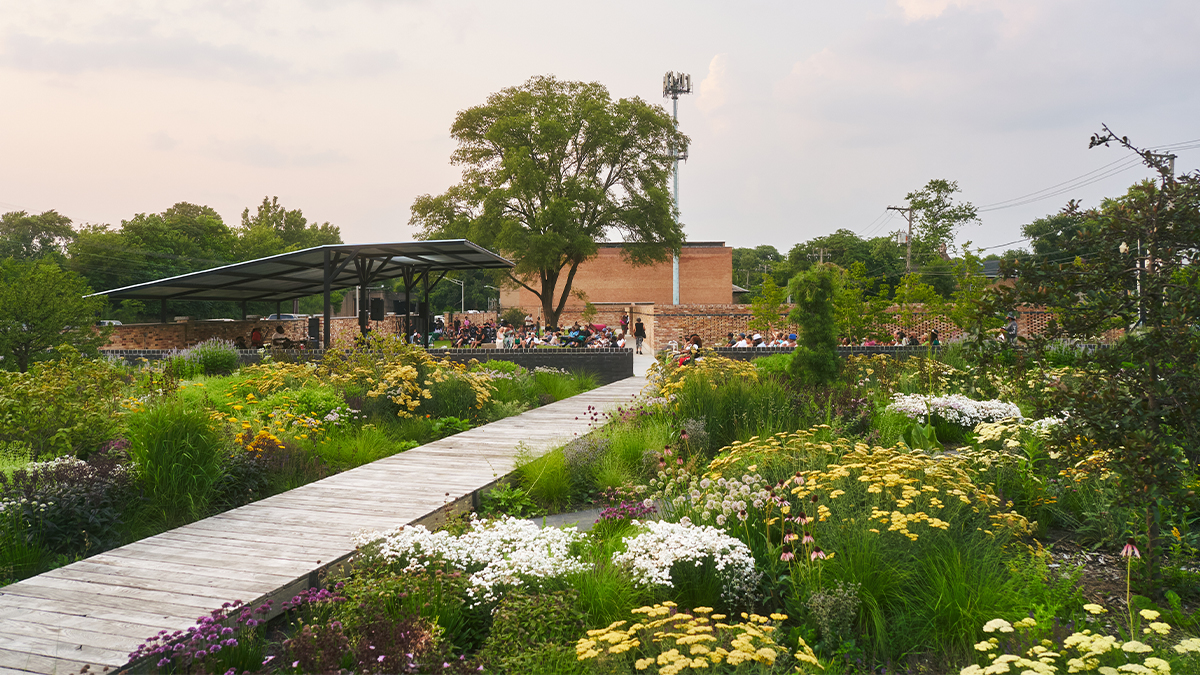 Boardwalk curves through garden toward pavilion in background; lush plants on either side.