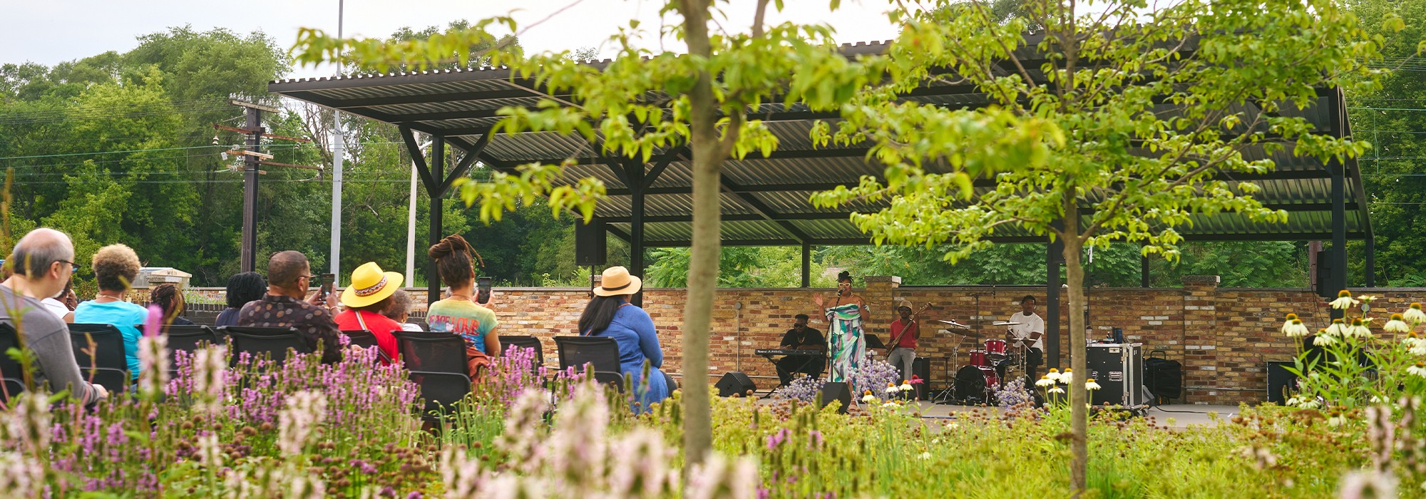 Flowering plants in foreground; seated people listening to a band under a pavilion.