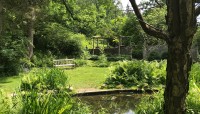 Pond in the foreground surrounded by trees and shrubs, wooden bench and gazebo in the background.