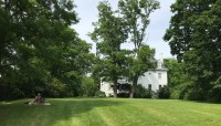 Lawn in the foreground with picnicking couple, mansion surrounded by shady trees in the background. 