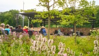 Flowering plants in foreground; seated people listening to a band under a pavilion.