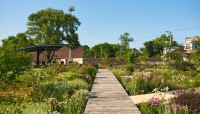 Boardwalk in the center flanked by flowering plants; pavilion on the left in the background.