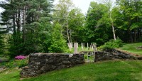 Stone wall enclosing outdoor rooms set in sloping lawn; trees in background