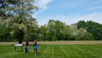 Three people walk in front of a field; trees in background