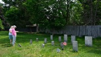Two women stand on the left; grave markers on the right; fence and trees in the background
