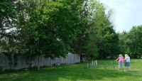 Two women stand looking at the grave markers on the right; lawn and fence extend to the left with trees
