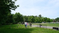 Two women stand looking at the grave markers on the left; road on the right with sports fields opposite