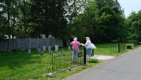 Street to the right lined by a fence; grave markers and two women on the left with a fence and trees in the background