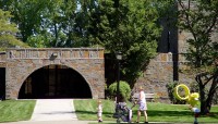 Family walks on sidewalk in front of an academic building with a tunnel entrance. Trees surround.