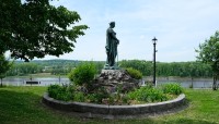 Statue in circular, raised bed; trees on either side; river and mountains in distance