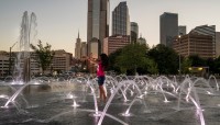 Girl jumps through water on splash pad; buildings in background