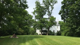 Lawn in the foreground with picnicking couple, mansion surrounded by shady trees in the background. 